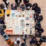 Students crowding around two instructors around a table which has lots of knick-knack items on it.