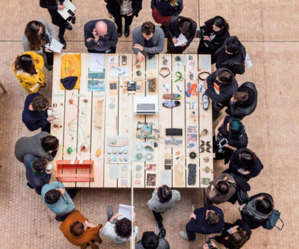 Students crowding around two instructors around a table which has lots of knick-knack items on it.