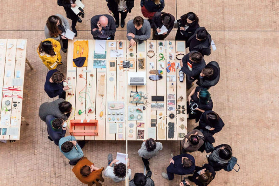 Students crowding around two instructors around a table which has lots of knick-knack items on it.